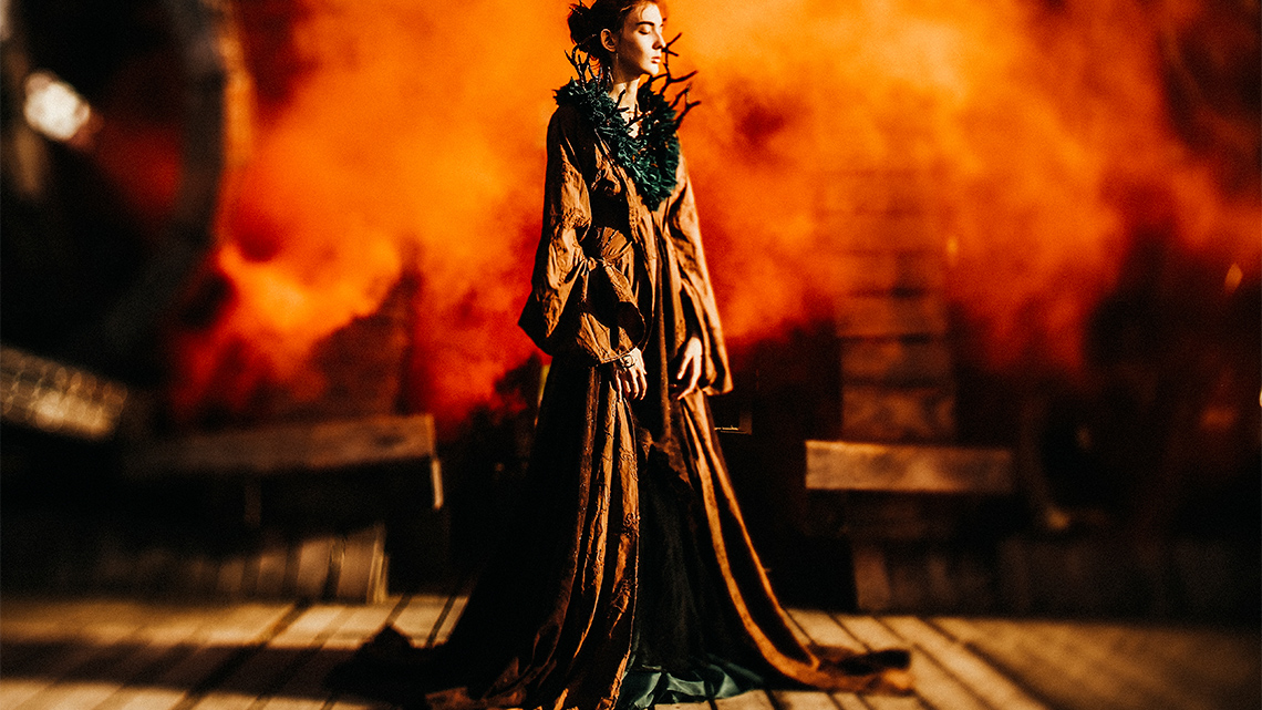 A person dressed in a flowing brown gown and a necklace made from branches stands in front of a fiery background.