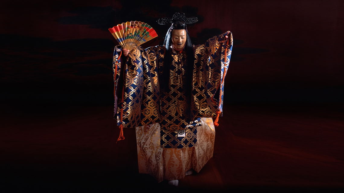 A person dressed in robes used in Noh theatre, holding a hand fan.