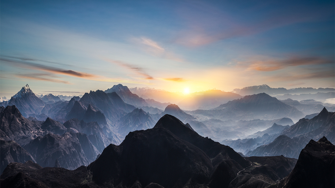 An aerial view of a misty mountain range at sunrise with thin clouds. A narrow road that winds between two mountains is at the bottom right.