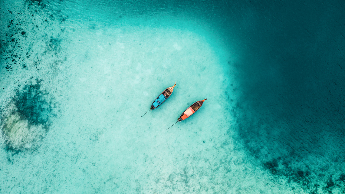 An aerial view that looks down on two boats that are floating on top of clear water. The sandy sea floor is visible underneath the boats.