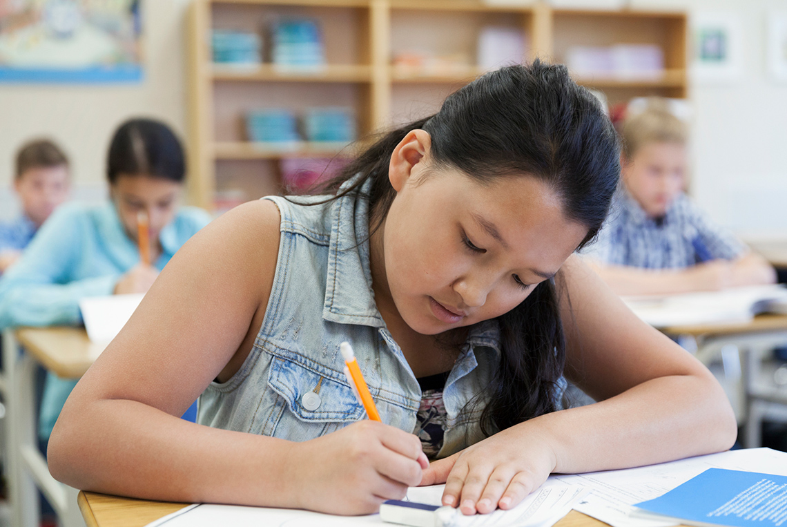 A student sits at a desk indoors in a classroom, writing on a piece of paper using a pencil, with an eraser and other papers beside them.