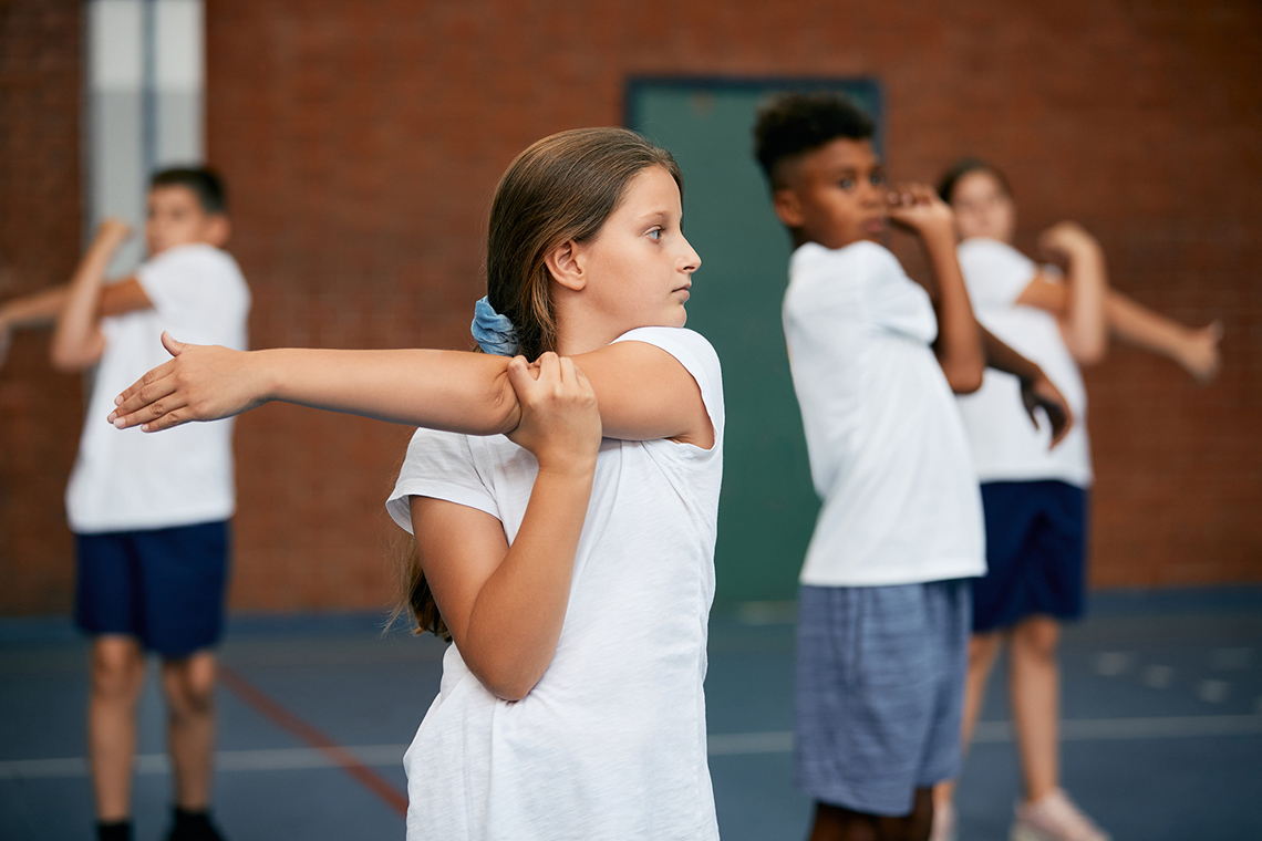 Warming up A student stands indoors in a gym, holding their left arm across their chest with their right hand, as they do stretches before gym class.