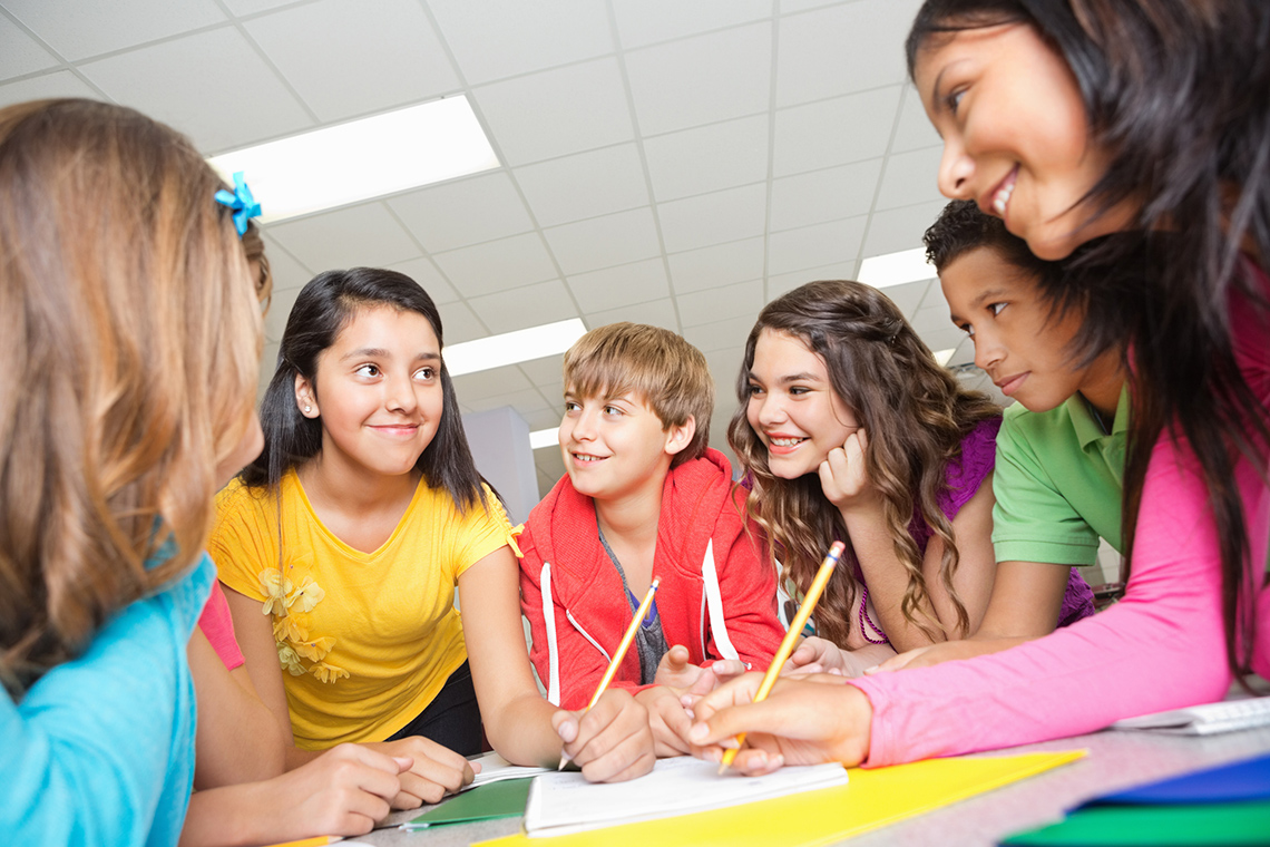 Group response Six students stand around a desk, leaning to look at 1 student in a yellow shirt. 2 students write with pencils in a notebook on the desk.