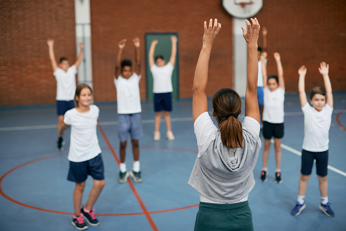 Warming up Six students stand indoors in a gym, all but 1 stretching their arms above their head, spaced out from each other, facing their teacher.