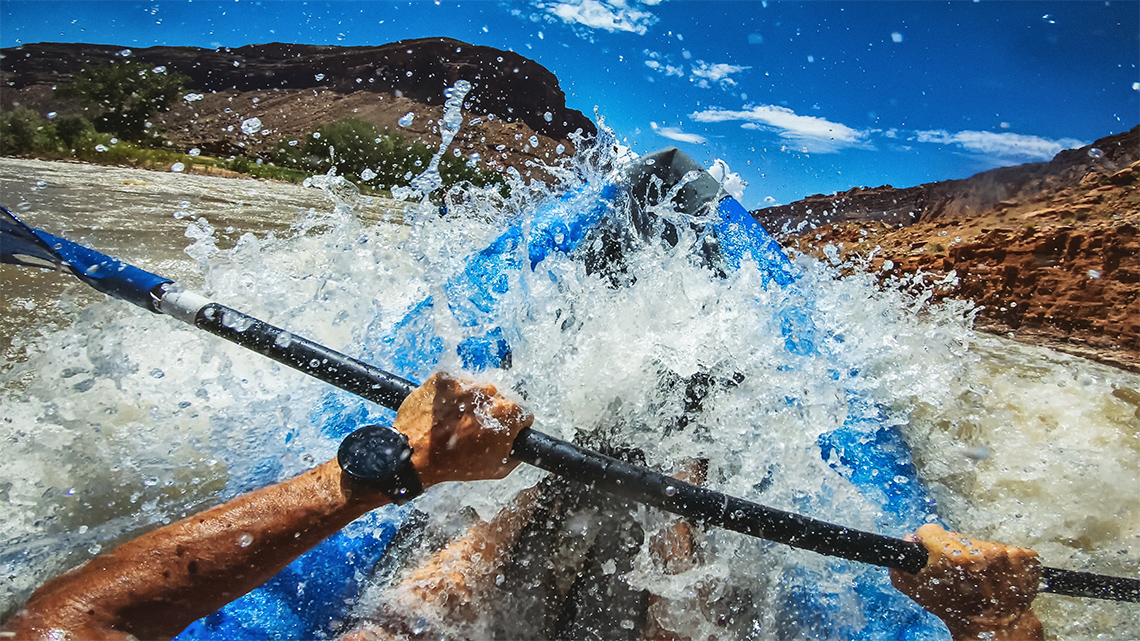 Perspective of a person navigating a kayak with a paddle through a fast-paced river surrounded by rocky hills on a clear sunny day.