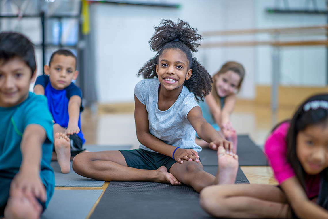 Students sit on yoga mats in rows on the floor of an indoor gym, 1 leg bent, the other straight out, stretching forward to touch their toes.