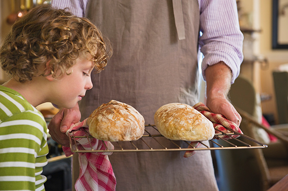 A student leans in to smell two freshly baked loaves of bread, which are sitting on a baking rack being held with two hands by an adult.