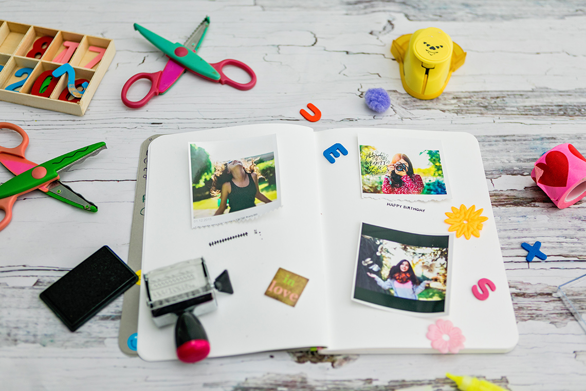 A scrapbook lies open on a desk, 3 photos lying on top of the pages, with an ink pad and stamp, scissors, letters, and other items.