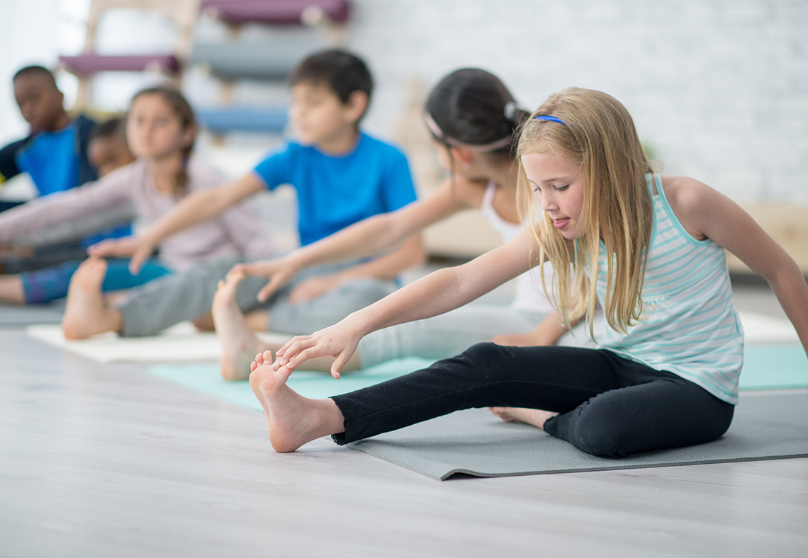 Students sit on yoga mats in a row on the floor of an indoor gym, 1 leg bent under the other leg which is straight out, touching their toes.