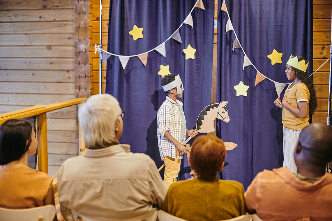 Two students stand in front of a curtain on a stage performing for 4 people. 1 student wears a crown, 1 wears a mask holding a hobby horse.