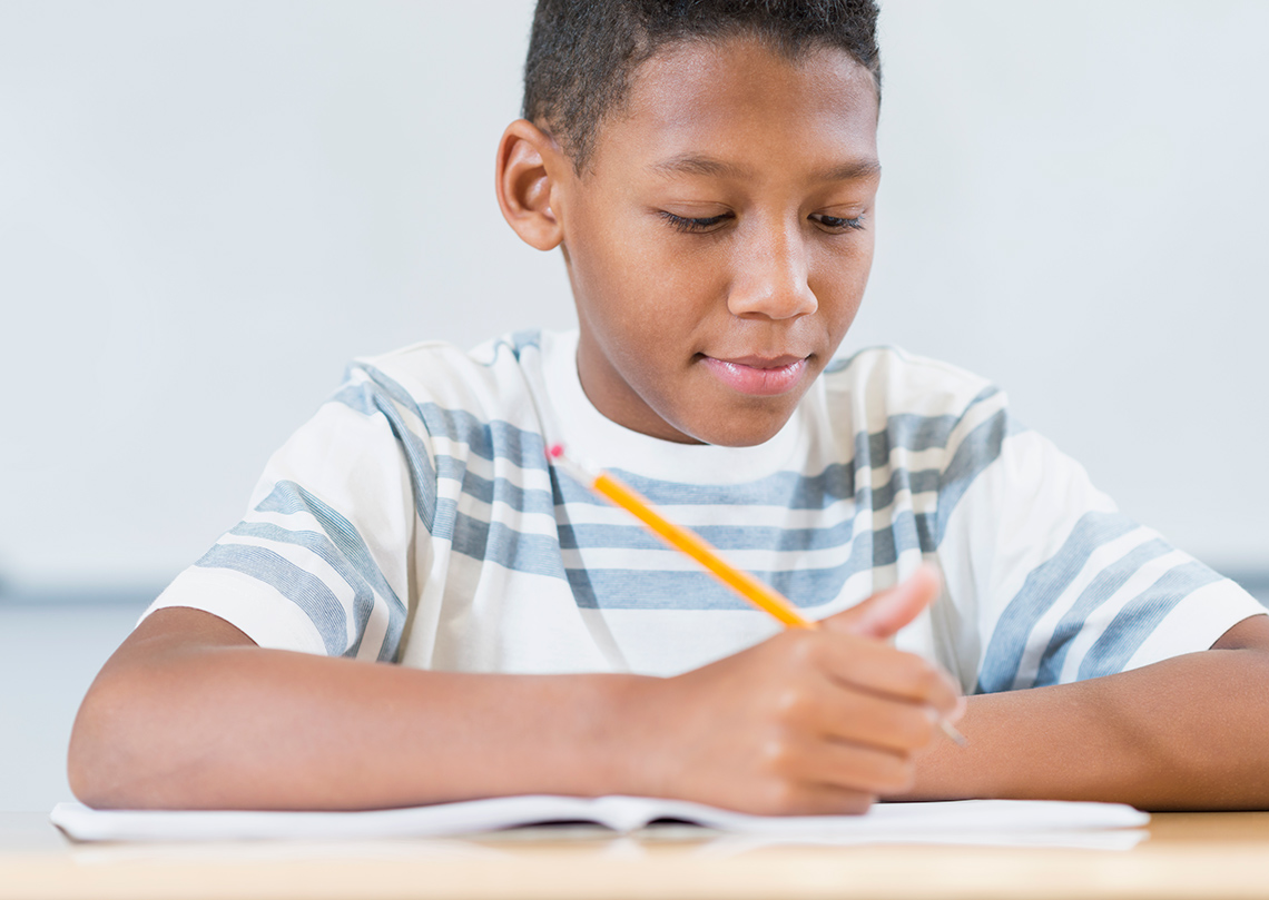 A student wearing a vertical-striped shirt sits at a desk indoors in a room, facing the camera, writing in an open notebook with a pencil.