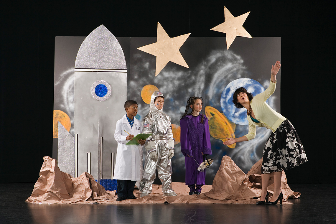 Three young actors on a space-themed stage, 1 in an astronaut suit, 1 in a lab coat, 1 wearing an alien mask listen to their stage director.