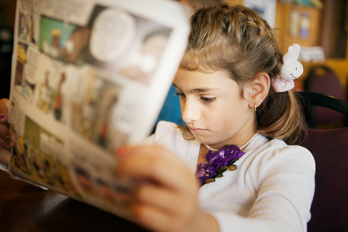 A student sits in a chair indoors holding up a page of a paper newspaper with comics printed on it as they read the other side of the paper.