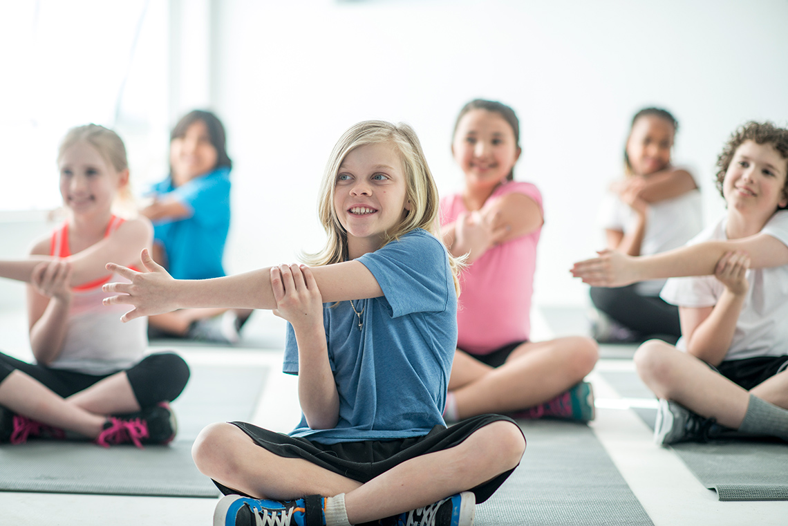 Students sit cross-legged on yoga mats in rows on a floor of an indoor gym, holding their left arm across their chest with their right hand.