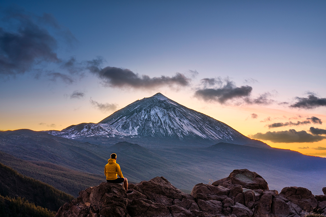 Person seated on a large rock while admiring the view of the volcano, Teide, at dusk, in Spain.