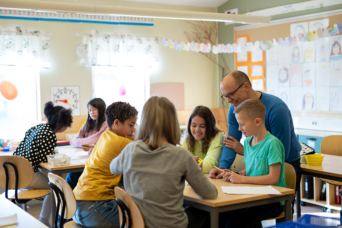 Students sit at 2 tables in a bright classroom, working together with paper and pens, a teacher stands behind 1 table to help the students.