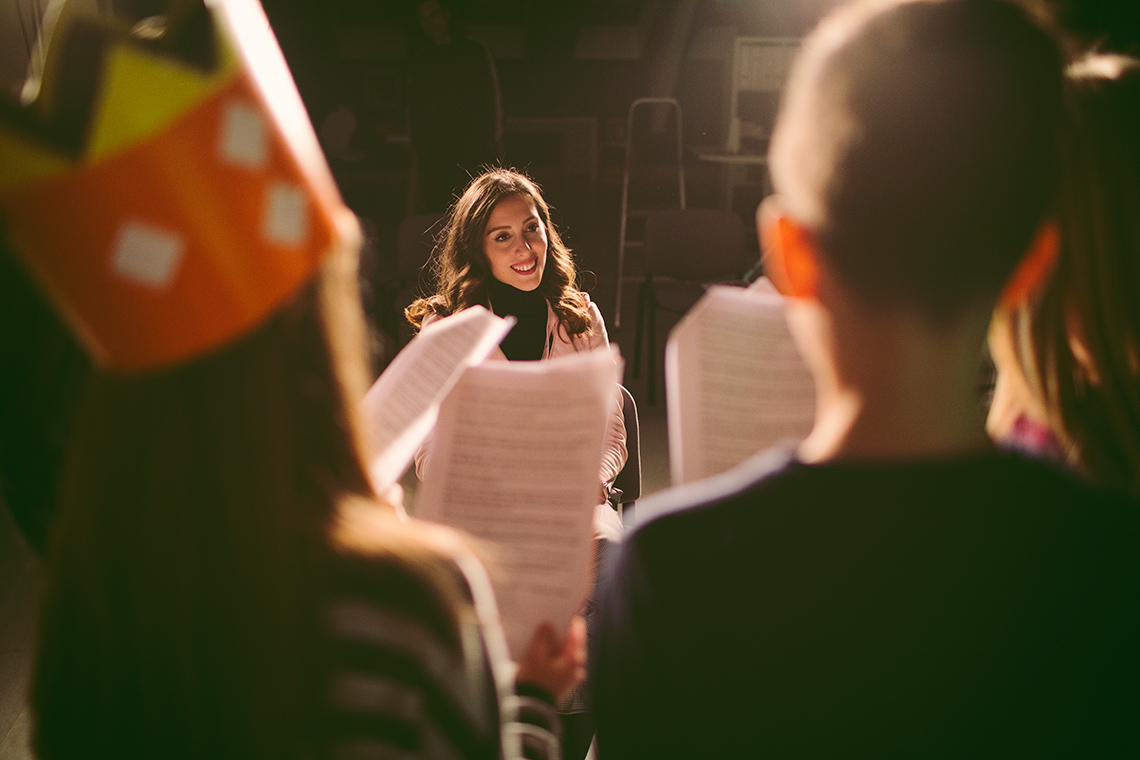 Students stand facing away from the camera holding up pieces of paper reading their lines in front of their teacher who stands on a stage.