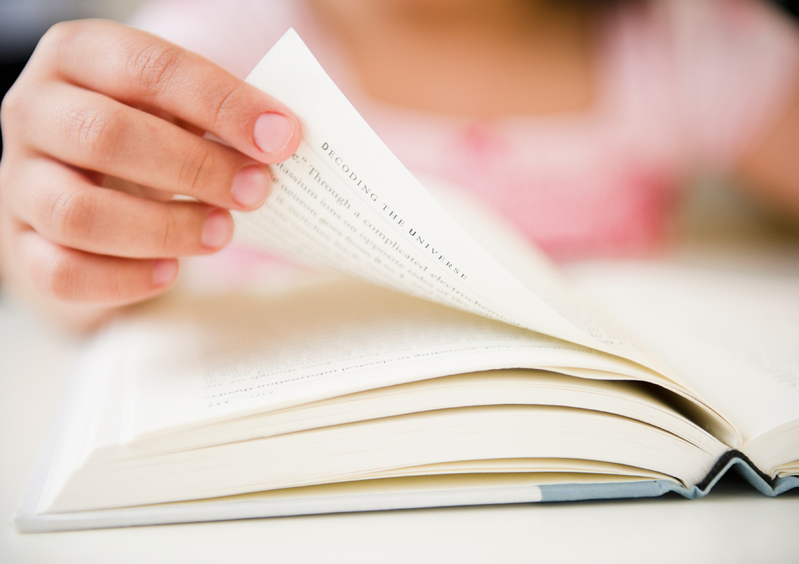 A student, blurred in the background, sits at a desk, a hardcover book open in front of them. They turn a page in the book with their hand.