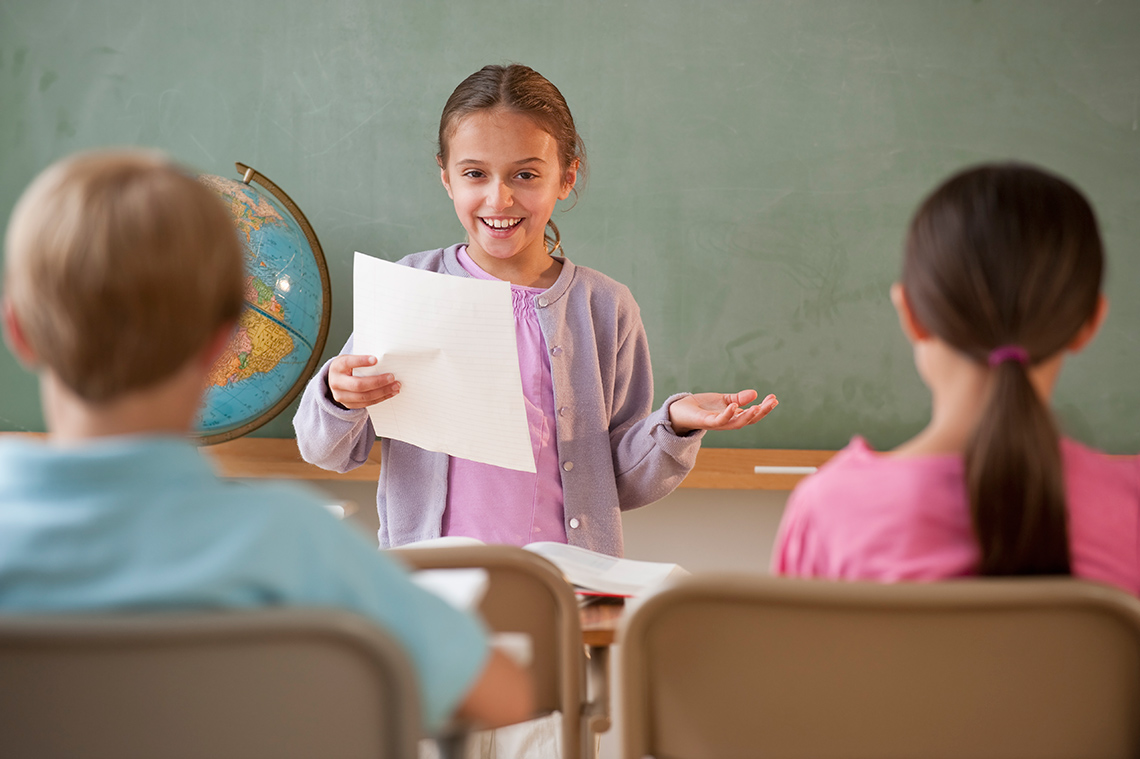 A student stands at the front of a classroom holding a piece of paper in 1 hand and gesturing with the other as they give a presentation.