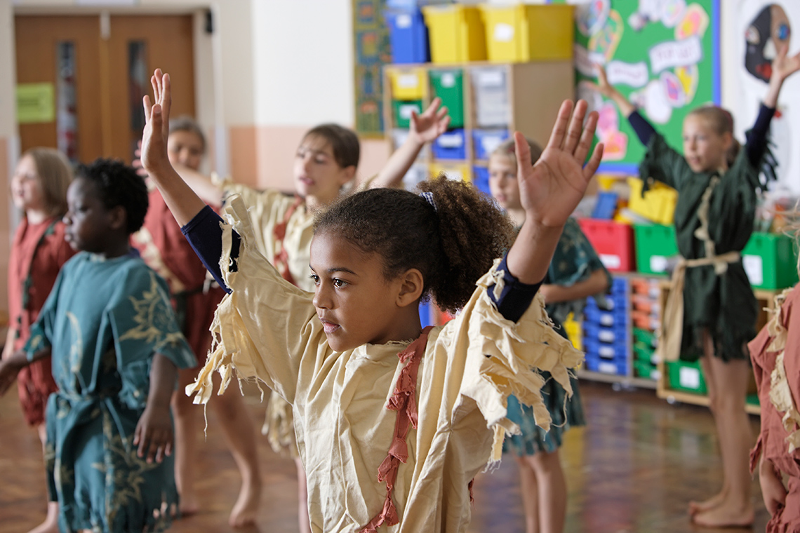 Practice Students stand in rows indoors in a classroom dressed in coloured tunics, some students have their arms raised up, perform in drama class.