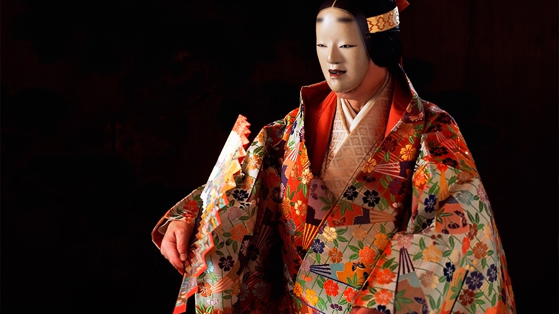 A performer in an elaborate Japanese Noh costume and holding a fan expresses sadness using body movements during a play.