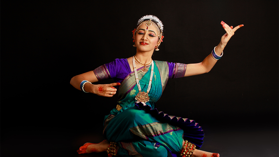 A Bharatanatyam dancer sits on the floor posing with one arm raised and the other arm at chest height. The dancer wears a colourful costume.
