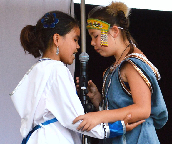 Inuit Throat Singing Two Inuit girls, dressed in traditional clothing, stand close together, arm in arm, holding a microphone and performing throat singing.