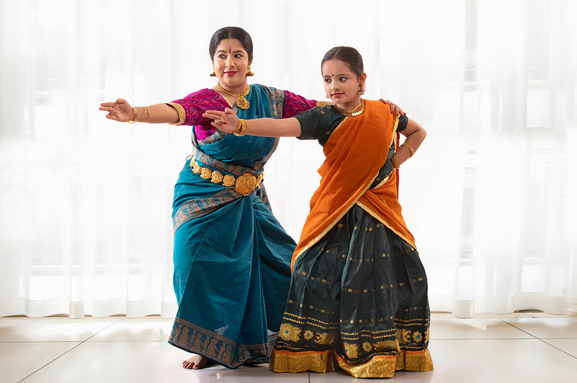 Bharatanatyam Bharatanatyam teacher stands behind a student, both dressed in colourful dresses, posing with their left arms pointing straight, legs bent.