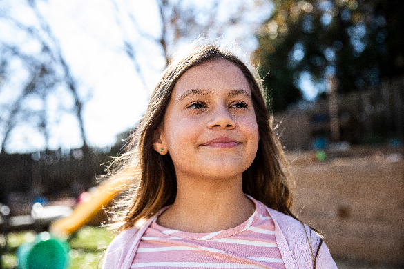 Child thinking A young person, wearing a t-shirt and sweater, stands outside in a playground on a sunny day, smiling and looking off to the right.