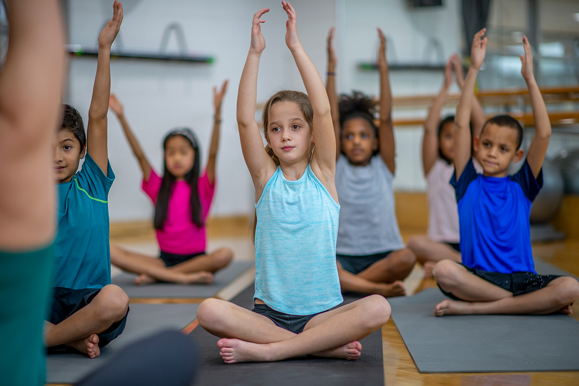 Stretching Arms Students sit cross-legged on yoga mats in rows on a floor of an indoor gym, both arms raised up above their heads, facing their teacher.