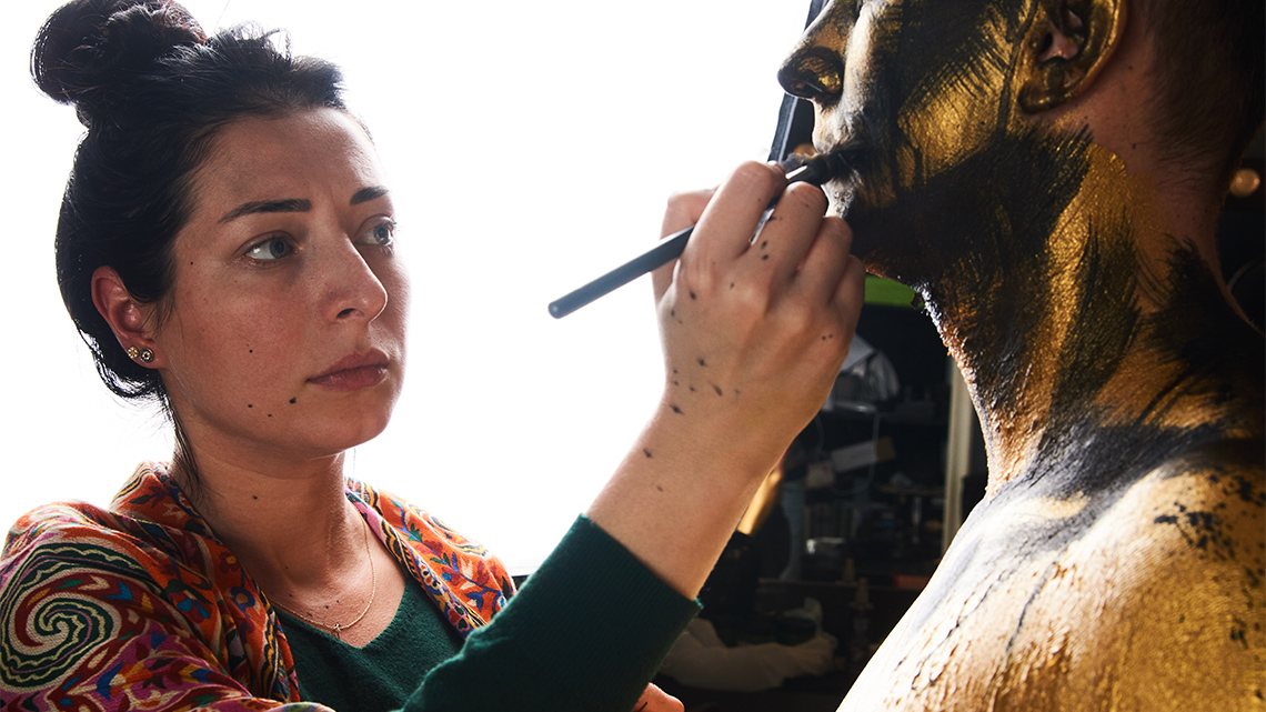 A makeup artist applies gold makeup, using a brush, on and around a performer’s mouth and face indoors in front of a bright light.