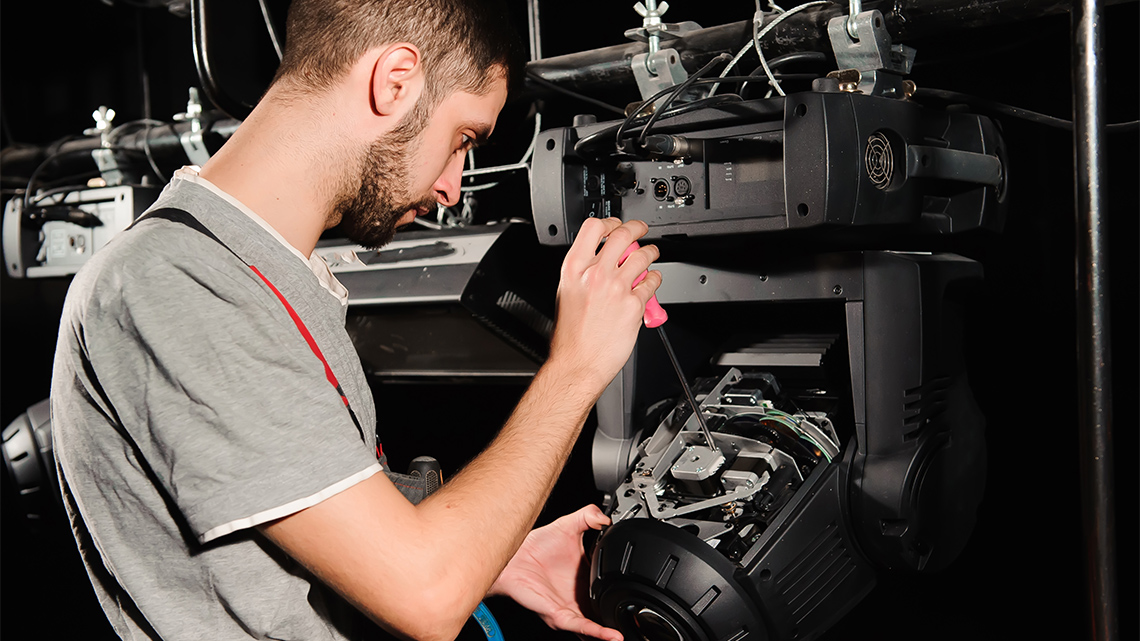 A lighting engineer or operator uses a screwdriver to adjust or fix the controls of a stage light indoors on, or behind, a stage.