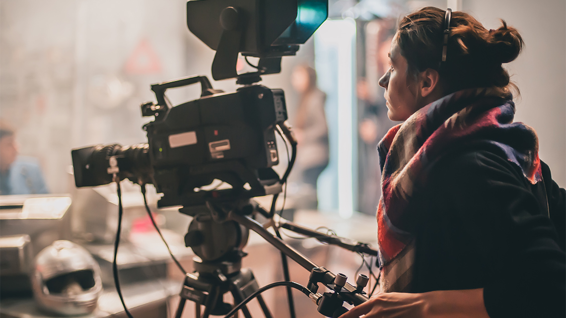 A camera person stands behind a movie camera, holding one handle with their left hand, controlling the camera while filming on a movie set.
