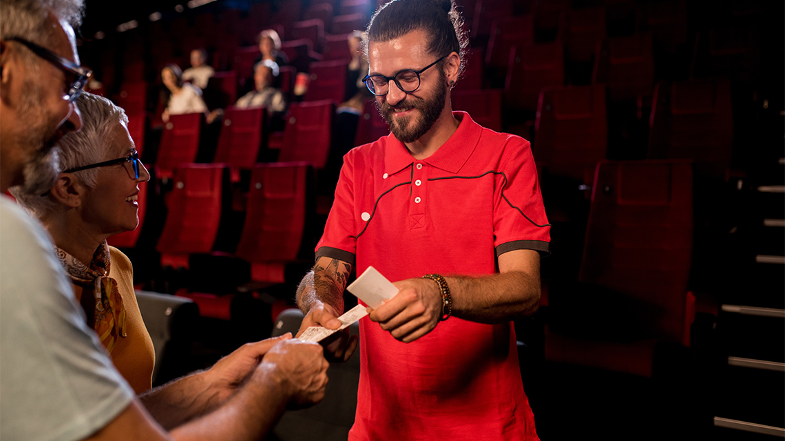 A theatre assistant stands on the steps of the outside aisle indoors in a theatre, as they check the tickets of two theatre attendees.