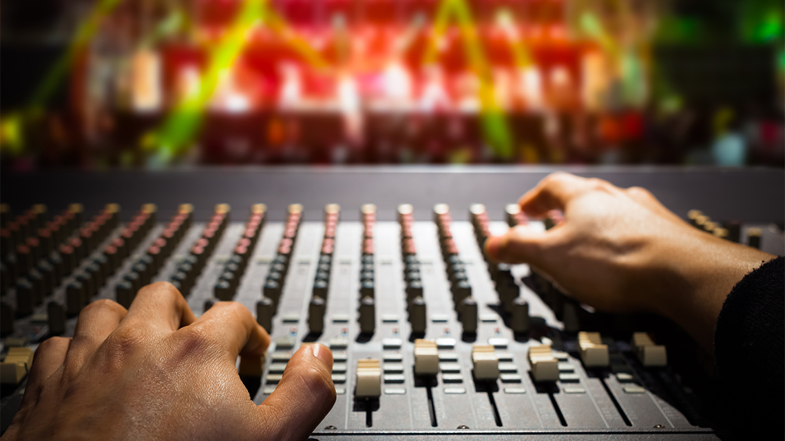 A close-up view of a sound engineer’s hands operating a sound mixer board for a concert being performed on a stage in the background.