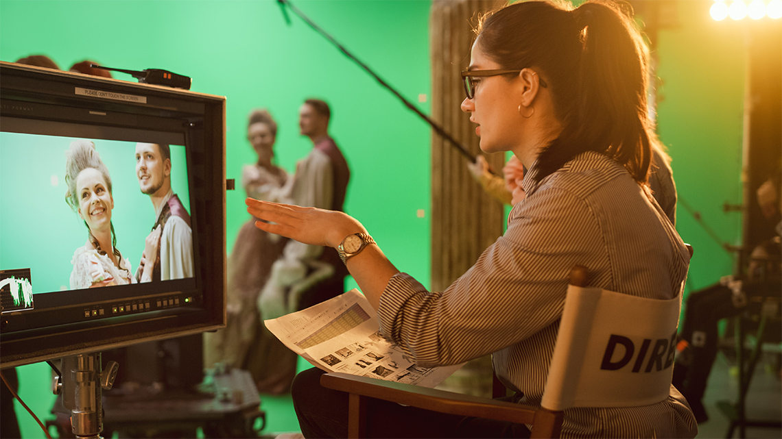 A director sits in a director’s chair looking at and pointing to a monitor showing actors in a film scene as they guide the crew and actors.