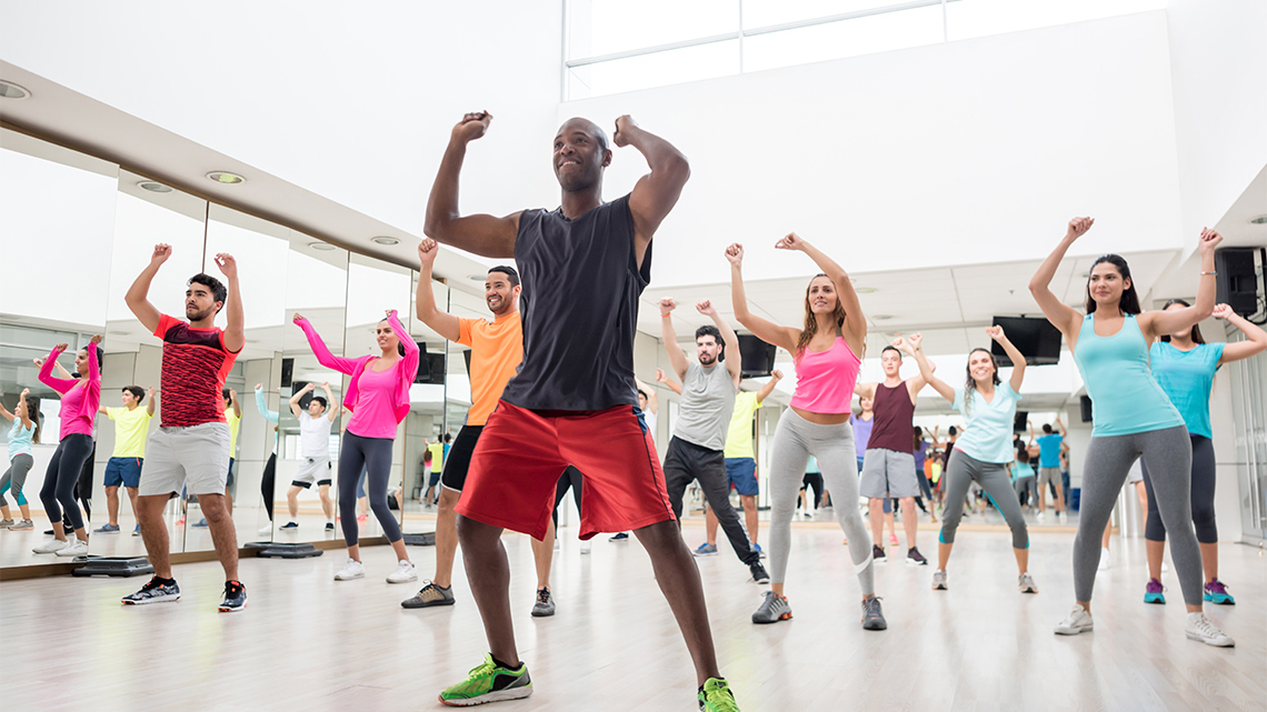 A choreographer dances in front of several dancers indoors in a dance studio, with mirrors on each of the walls, as they lead a dance class.