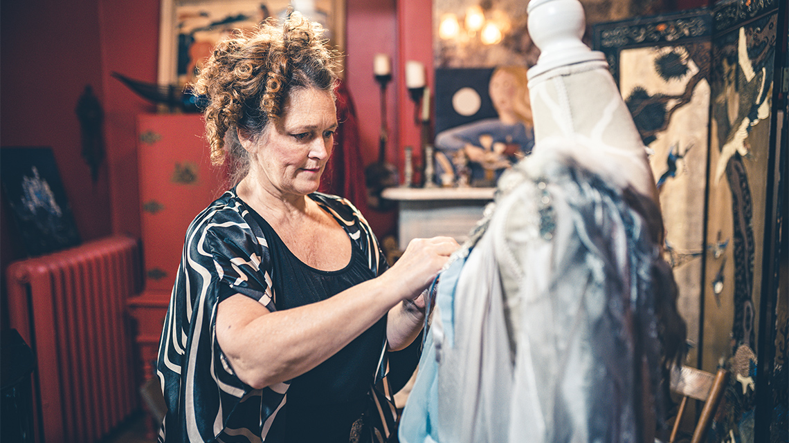 A costume designer makes alterations or adjustments to an outfit that is on a mannequin indoors in a prop room or on a movie set.