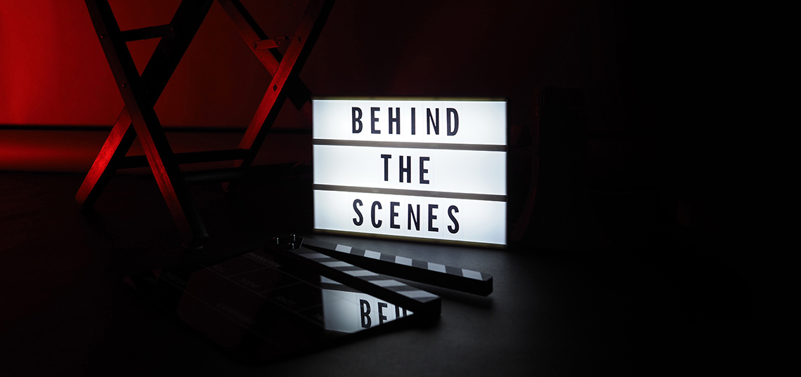Behind the scenes A lightbox with the words Behind the Scenes on it sits on a dark stage beside a director’s chair and behind a movie slate clapperboard.