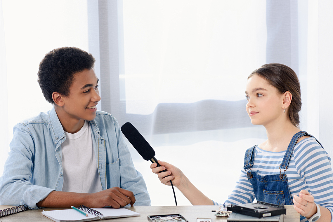 Interview time Two students sit at a table indoors in a bright room, one student holds a microphone out to the other student as they conduct an interview.