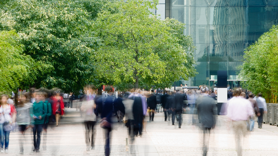 There are people quickly moving around on a concrete walkway, heading toward or away from a large, glass building.