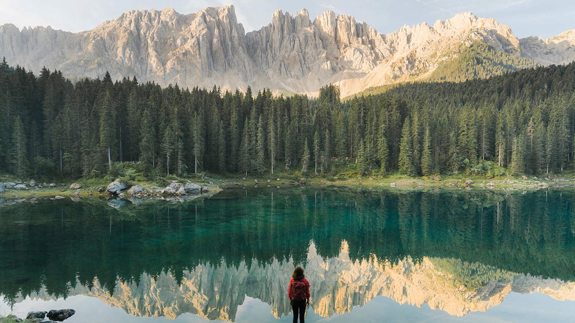 A person standing on a rock in front of a still lake. There is a forest of tress and mountains in the background.