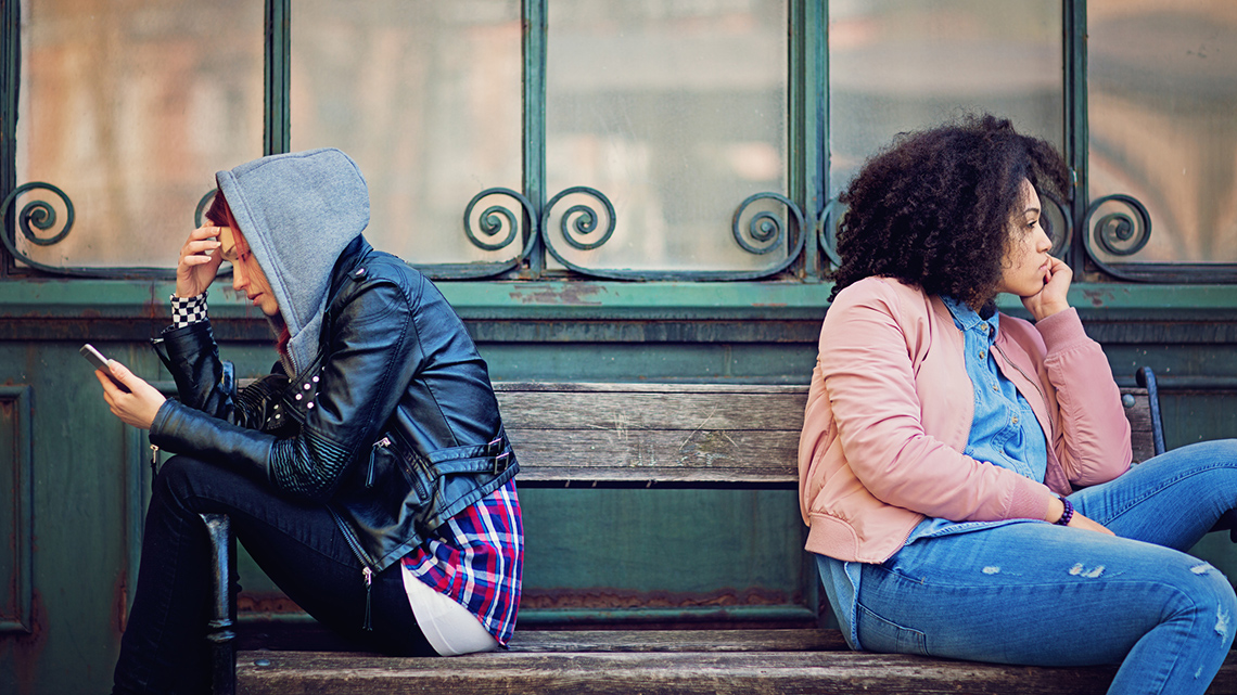 Two female teens sitting on opposite ends of a bench with their backs toward one another
