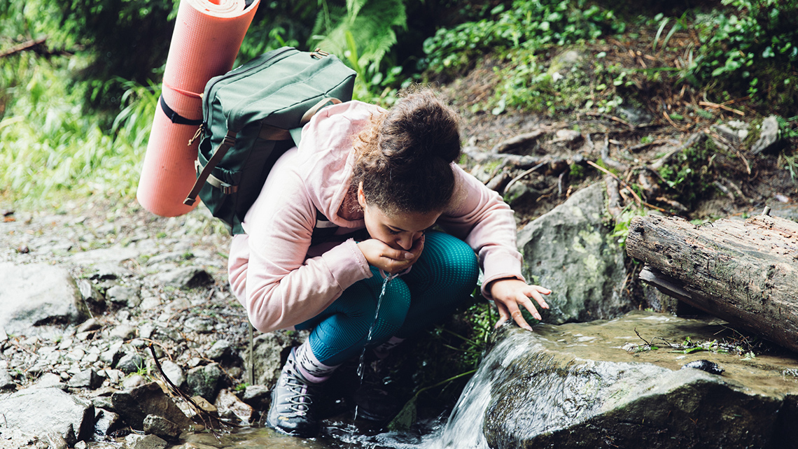 A youth stops to drink from a running stream of water. They are carrying a backpack with a yoga mat.