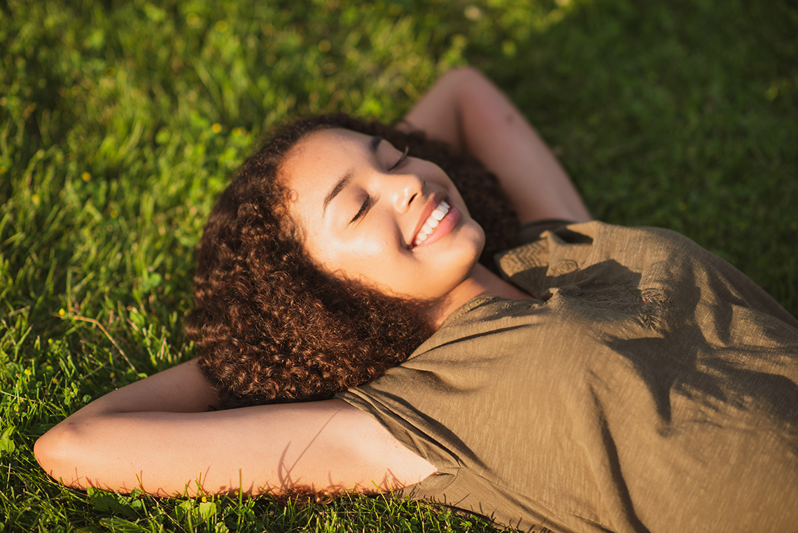 A young person lies on their back on the grass outdoors on a sunny day, their arms placed underneath their head, eyes closed and smiling.
