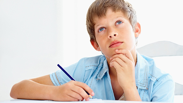 A young person sits at a desk indoors, holding a pencil, with their hand under their chin, looking up towards the ceiling in deep thought.