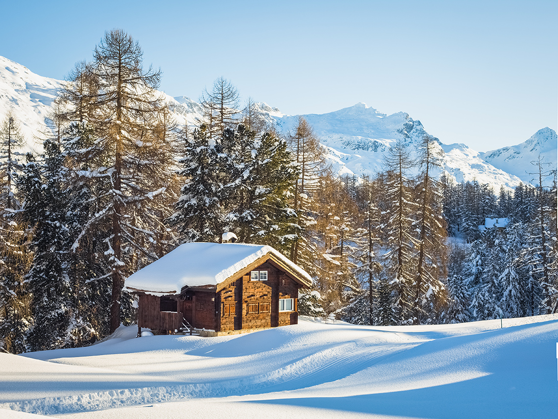 A log cabin with a snow-covered roof sits in front of a row of trees, mountains in the background, snow drifts in front on a cold sunny day.