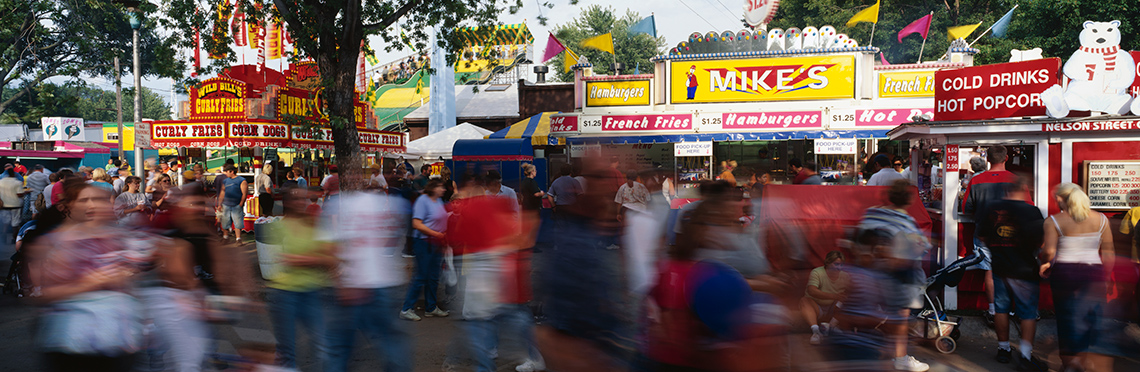 A crowd of people walk past colourful food stands on a midway at a local fair on a sunny day. The people in the foreground are blurred.