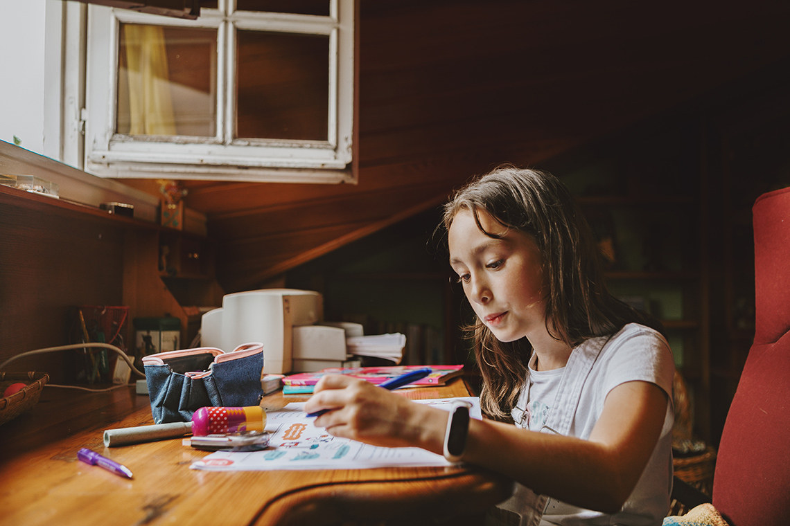 A young person sits at a table indoors, drawing on a piece of paper with a pen, with an open pencil case, markers, and pens on the table.