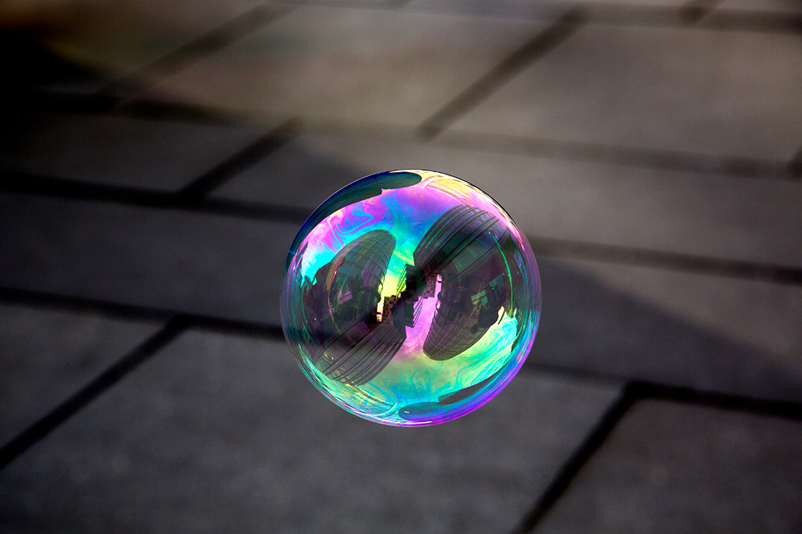 A large soap bubble floats in the air outdoors above a sidewalk or patio. The bubble shows a distorted reflection of some buildings nearby.